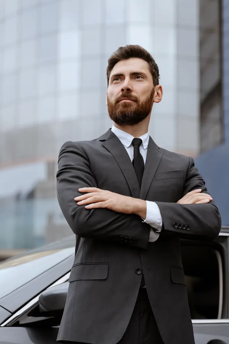 Professional chauffeur in grey suit standing confidently beside luxury car in front of modern glass building