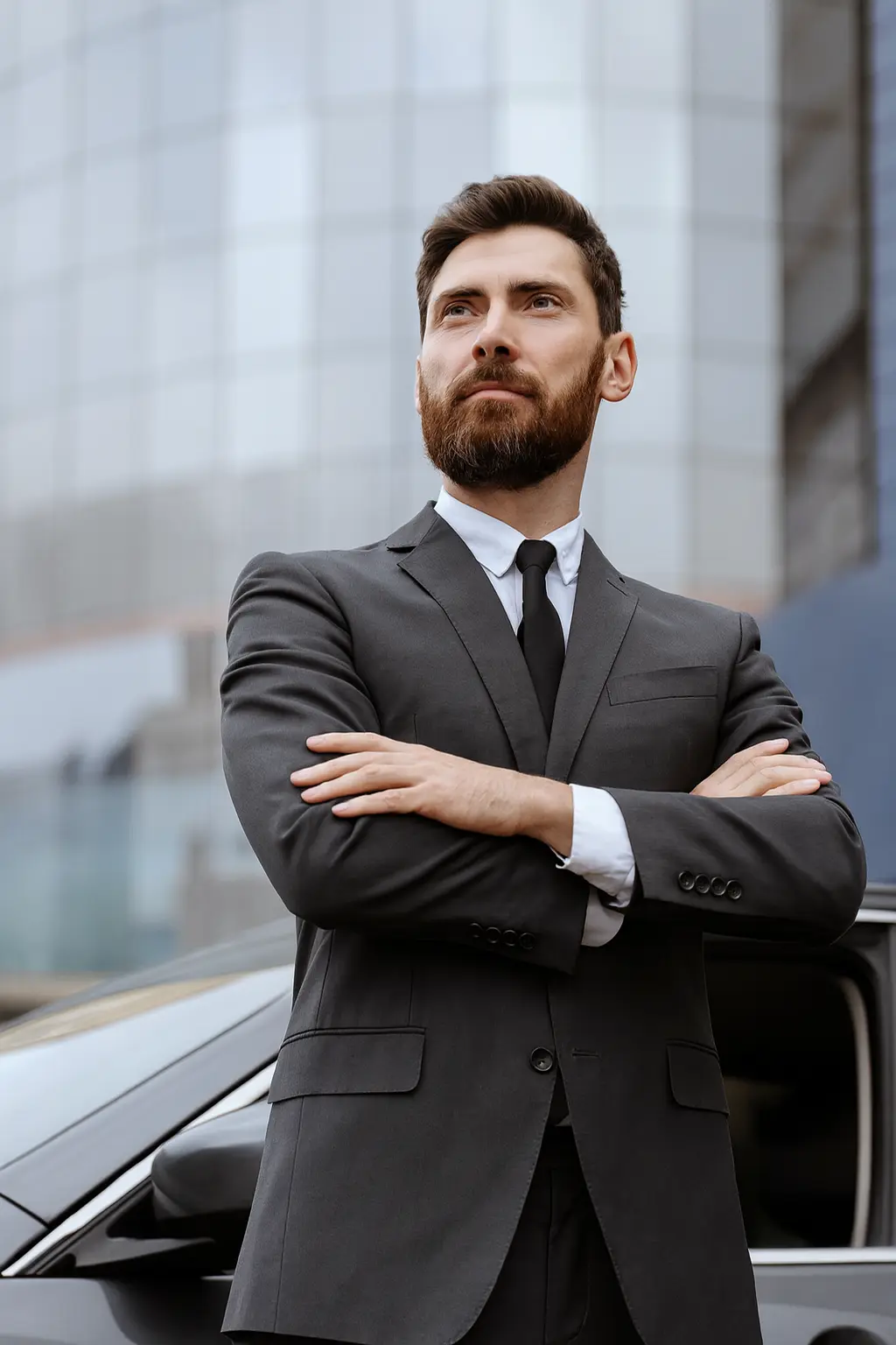 Professional chauffeur in grey suit standing confidently beside luxury car in front of modern glass building
